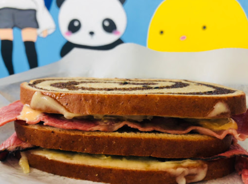 A layered sandwich with meat and cheese on swirled bread, set against a colorful cartoon background.